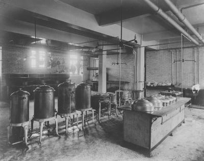 Black and white image of Ryan's kitchen in 1906 showing tiled walls lined with pots and pans and several large cooking vats in the center of the room. 