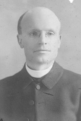 Black and white headshot of Fr. David Buel wearing a clerical collar under a black overcoat.