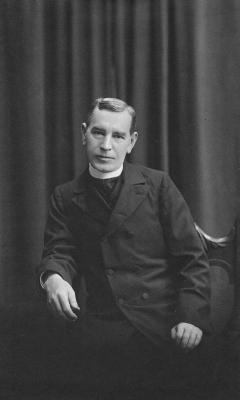 Black and white photo of Fr. Jerome Daugherty seated in a wooden chair wearing a clerical collar underneath a black overcoat.