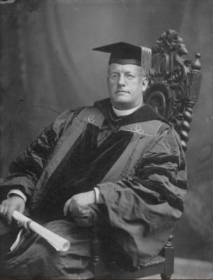 Black and white image of Fr. John D. Whitney seated in a wooden chair, clothed in academic regalia complete with cap and a diploma in one hand. 