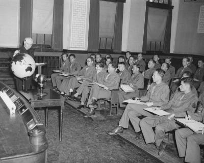 Fr. Walsh gestures at a globe as he lectures a class of uniformed students in 1945