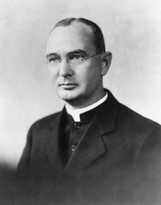 Black and white headshot of Fr. Arthur O'Leary wearing clerical robes underneath a black overcoat.