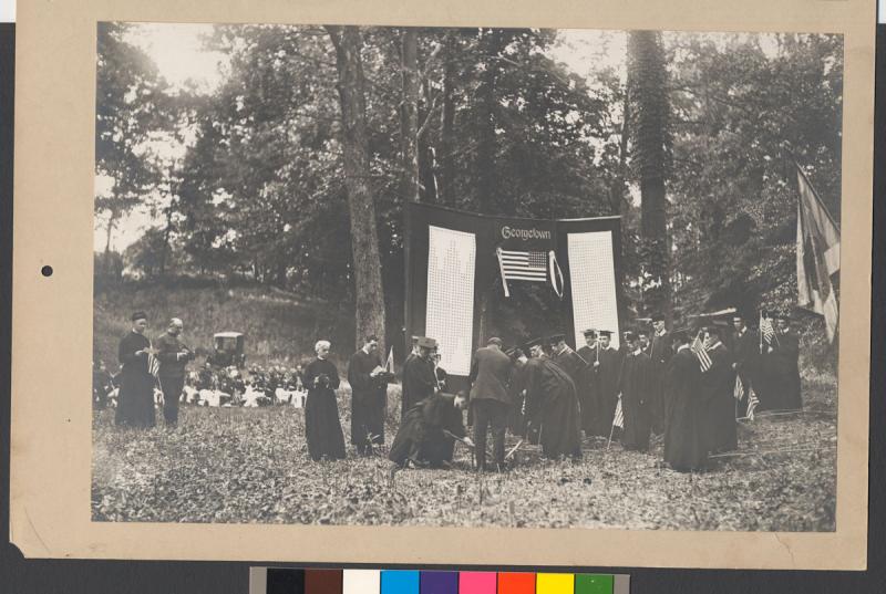 Black and white photo of Georgetown seniors in commencement caps and gowns solemnly planting trees under a Georgetown banner.