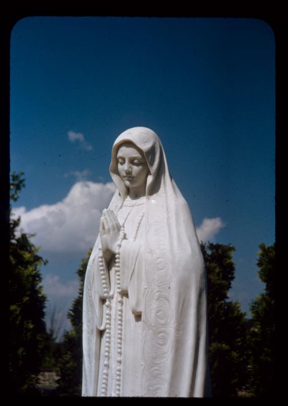 Photo of the marble statute of Our Lady of Fatima who is depicted with hands clasped and head bowed in front of a bright blue sky.